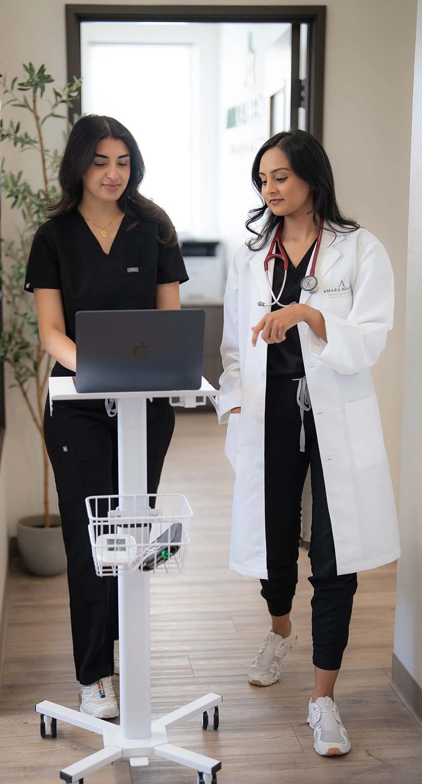 Two Amara Health clinicians reviewing notes on a laptop cart in the corridor between exam rooms.