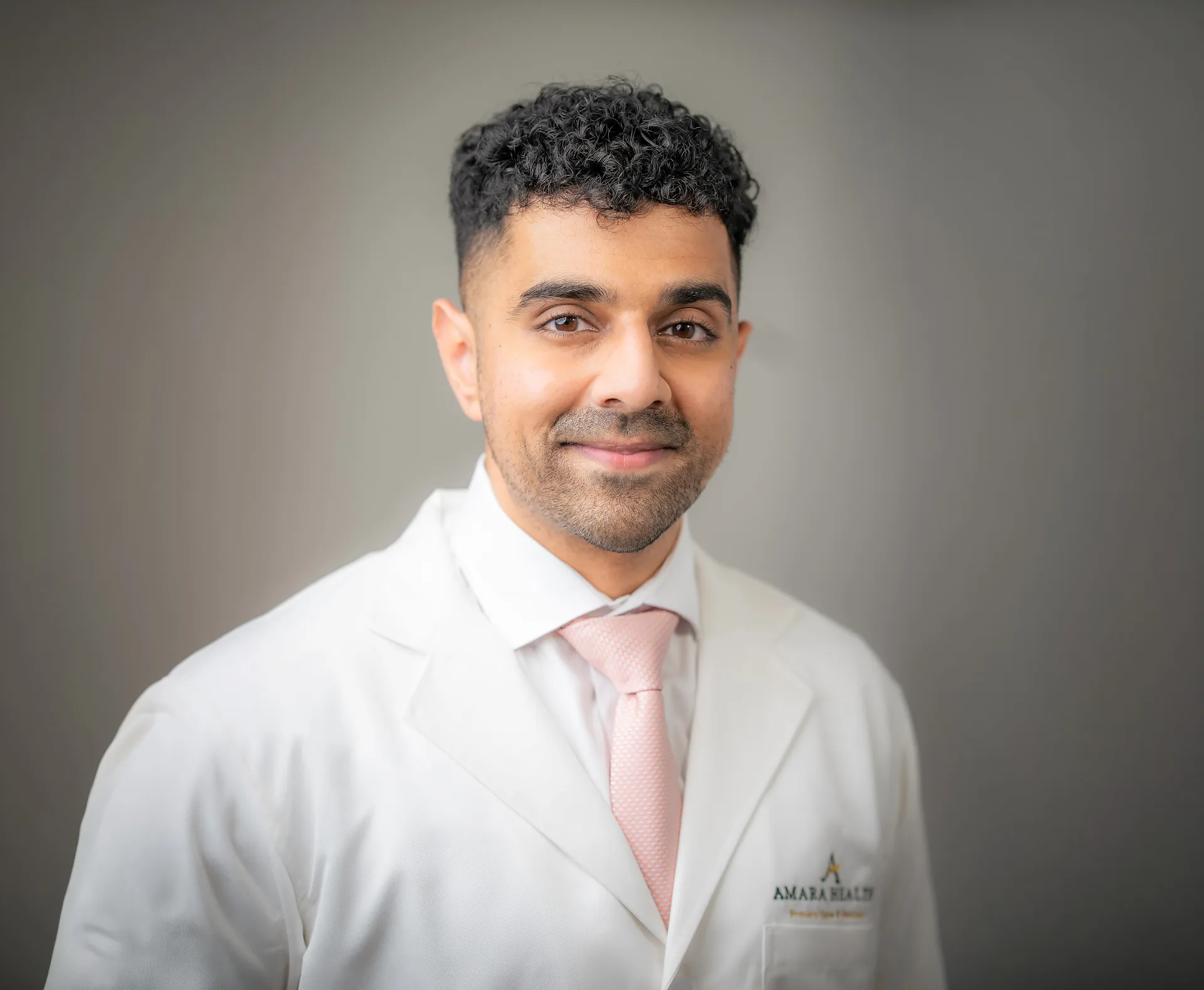 Dr. Krishna Ramesh in a white coat and tie, photographed against a plain gray background.