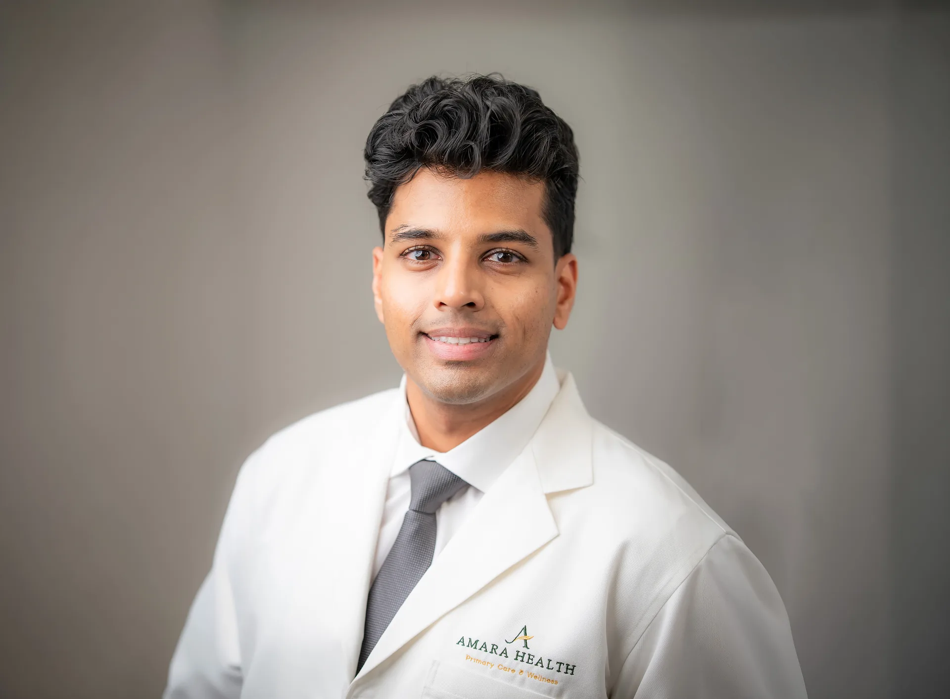Dr. Rangish Yuvaraj in a white coat and gray tie, photographed against a plain gray background.